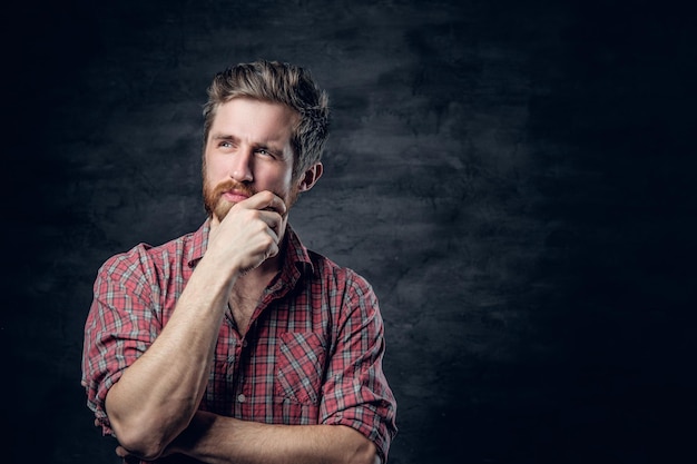 Retrato de estúdio de homem barbudo loiro vestido com uma camisa de lã vermelha sobre fundo cinza escuro.