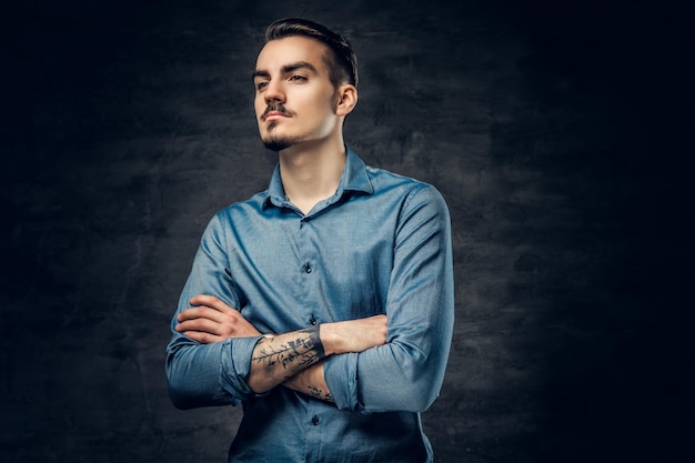 Retrato de estúdio de homem elegante com braços cruzados, vestido com uma camisa azul sobre fundo cinza.