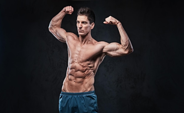 Retrato de estúdio de homem sem camisa de músculo ectomorfo mostra seu bíceps sobre fundo cinza escuro.