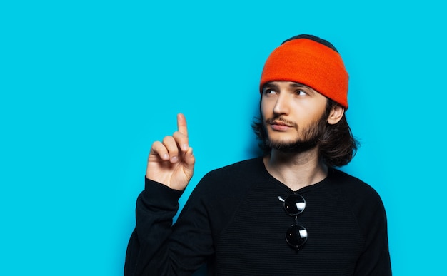 Retrato de estúdio de jovem apontando para longe contra um fundo azul. Usando um gorro laranja e um suéter preto.