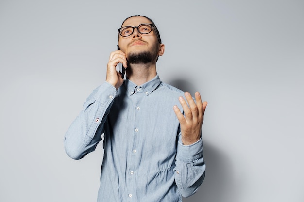 Retrato de estúdio de jovem falando no smartphone em fundo branco, vestindo camisa azul e óculos, olhando para cima e gesticulando com a mão