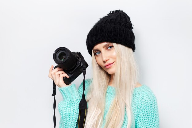 Retrato de estúdio de jovem loira segurando a câmera fotográfica, vestindo blusa azul e chapéu preto sobre fundo branco.