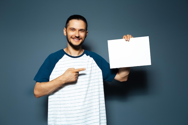 Retrato de estúdio de jovem sorridente apontando o dedo na placa de papel vazia em outra mão no fundo de cor azul