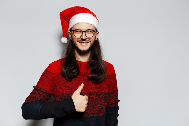 Retrato de estúdio de jovem sorridente com cabelos longos e óculos mostrando polegares para cima usa suéter vermelho de Natal e chapéu de Papai Noel em um fundo branco