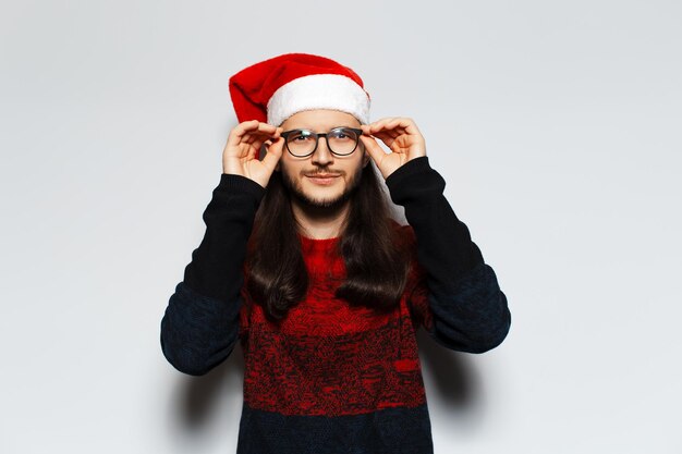 Retrato de estúdio de jovem sorridente com cabelos longos e óculos tocando óculos usa suéter vermelho de Natal e chapéu de Papai Noel em um fundo branco