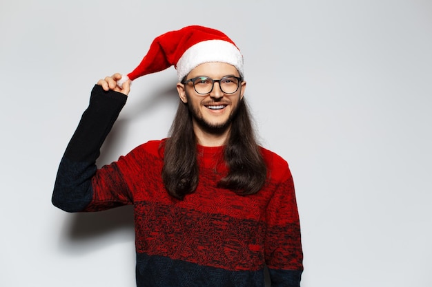 Retrato de estúdio de jovem sorridente com cabelos longos e óculos usa suéter vermelho de Natal segurando pompon de chapéu de Papai Noel em um fundo branco