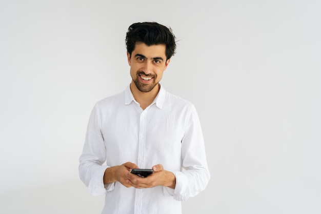 Retrato de estúdio de jovem sorridente em camisa casual olhando para câmera segurando smartphone usando aplicativos móveis online