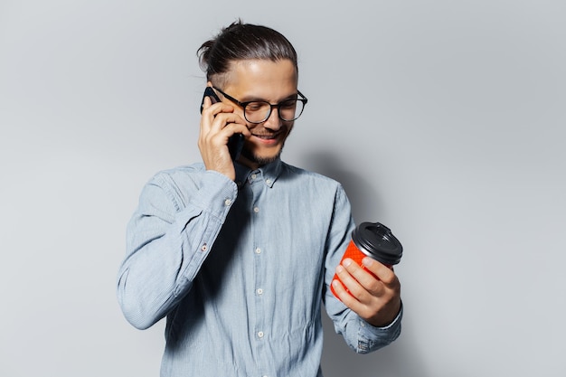 Retrato de estúdio de jovem sorridente falando no smartphone segurando um copo de papel vermelho para viagem de café em outra mão no fundo branco vestindo óculos e camisa azul