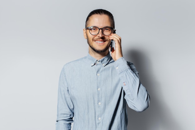 Retrato de estúdio de jovem sorridente falando no smartphone usando óculos e camisa azul sobre fundo cinza