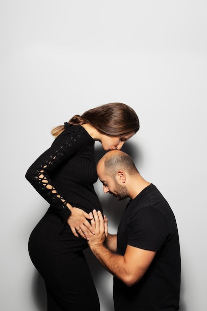 Retrato de estúdio de jovens futuros pais grávida vestindo vestido preto beija a cabeça de seu homem que toca a barriga no fundo branco