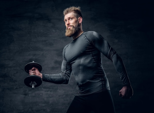 Retrato de estúdio de macho barbudo desportivo vestido com um sportswear cinza detém haltere.