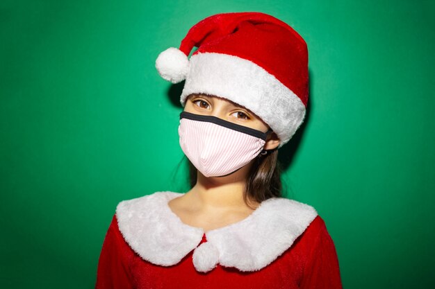 Retrato de estúdio de menina criança fantasiada de papai noel com chapéu vermelho e máscara médica na superfície verde