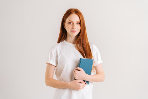 Retrato de estúdio de mulher jovem estudante sorridente segurando no livro de papel de mãos e olhando para a câmera, de pé sobre fundo branco isolado. Senhora ruiva feliz posando com materiais educacionais.