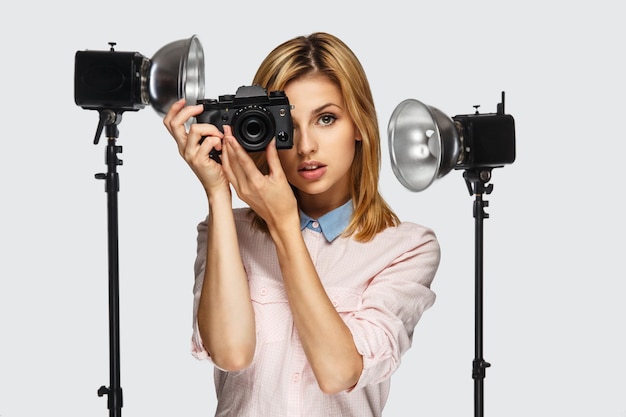 Retrato de estúdio de mulher loira positiva com câmera fotográfica com equipamento fotográfico no fundo.