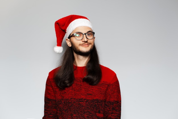 Retrato de estúdio de um jovem engraçado com cabelos longos e óculos usa suéter vermelho de Natal e chapéu de Papai Noel em um fundo branco