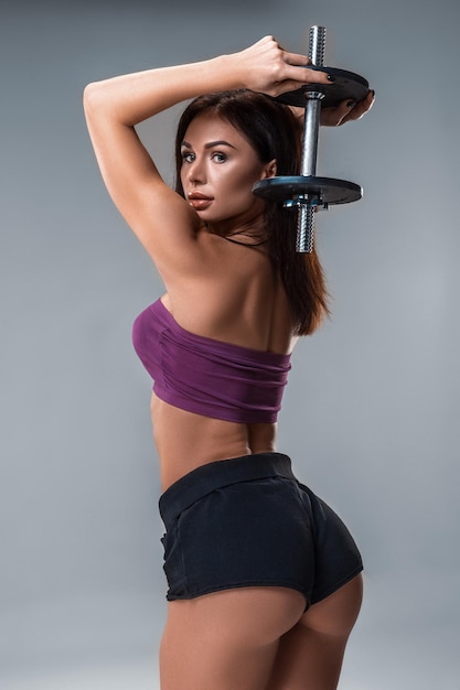 Retrato de estúdio de uma jovem desportiva posando com halteres contra um fundo cinza. Uma mulher está vestida com shorts curtos em um top roxo. Big Booty. Fechar-se