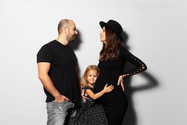Retrato de estúdio de uma jovem família feliz mãe e pai olhando cada um enquanto a filha abraçando a barriga no fundo branco usando chapéu vestido de preto
