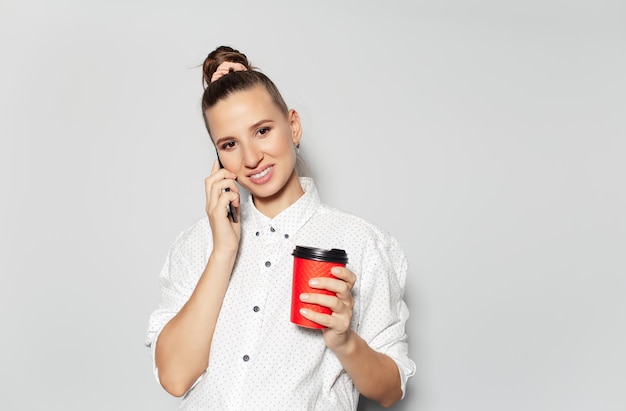 Retrato de estúdio de uma jovem linda falando no smartphone segurando uma xícara de café de papel em fundo branco