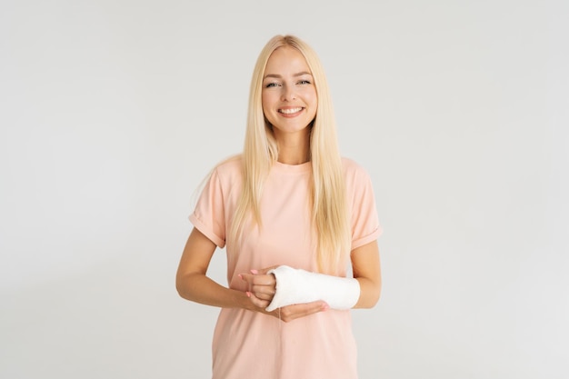 Retrato de estúdio de uma jovem loira alegre com o braço quebrado envolto em bandagem de gesso sorrindo olhando para a câmera em pé sobre fundo branco isolado