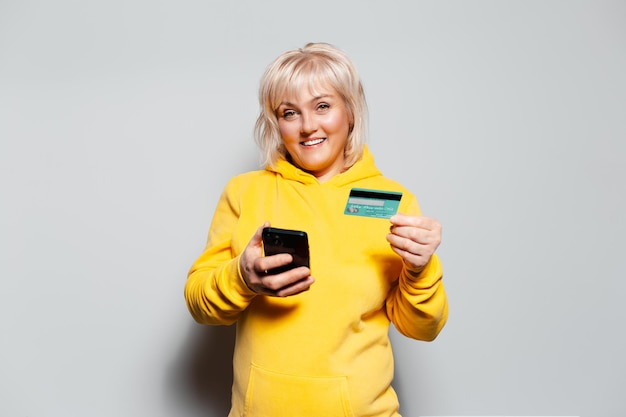 Retrato de estúdio de uma loira feliz com um suéter de capuz amarelo segurando um smartphone e um cartão de crédito nas mãos sobre fundo branco