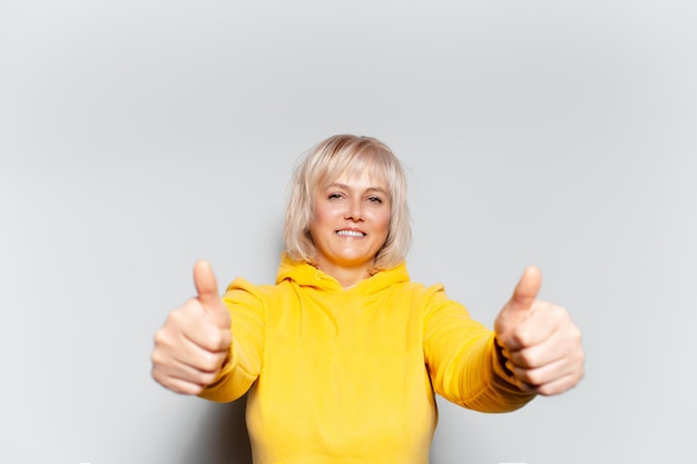 Retrato de estúdio de uma loira feliz mostrando os polegares para cima no fundo branco usando um suéter amarelo