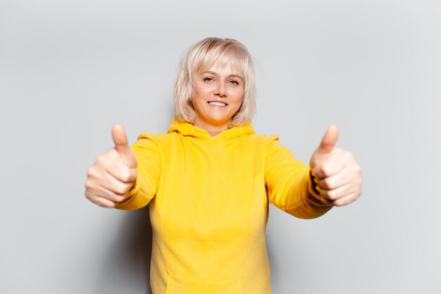 Retrato de estúdio de uma loira feliz mostrando os polegares para cima no fundo branco usando um suéter amarelo