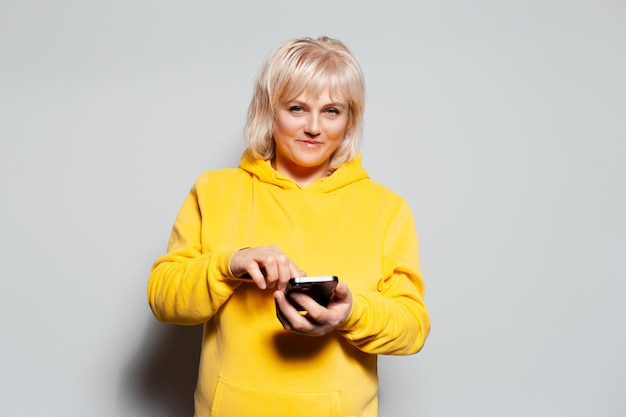 Retrato de estúdio de uma mulher loira de suéter amarelo usando smartphone em fundo branco