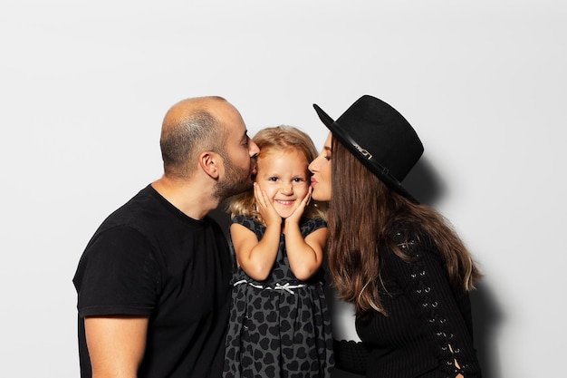 Retrato de estúdio do jovem pai de família feliz e mãe grávida beija sua filha em fundo branco usando chapéu vestido de preto