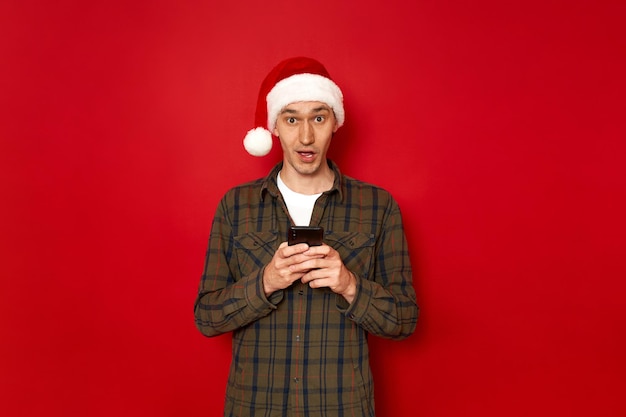 Retrato de estúdio horizontal de chocado celebrando homem segurando um telefone celular com roupas casuais e chapéu de Natal