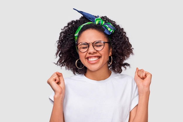 Foto retrato de estúdio horizontal de jovem com olhos fechados, punhos cerrados, alegra-se com o sucesso usa camiseta branca e faixa verde usando óculos redondos transparentes sobre fundo branco