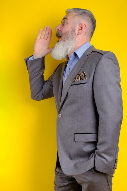Retrato de estúdio maduro homem barbudo em terno cinza grita e chama à parte, pesquisa e chamada de conceito, fundo amarelo.