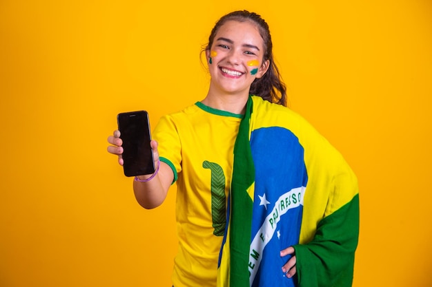 Retrato de fã brasileiro fã brasileiro mostrando seu celular vestido de fã de futebol ou jogo de futebol em fundo amarelo cores do brasilcopa do mundo
