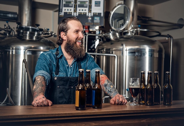 Retrato de fabricante masculino hipster tatuado e barbudo apresentando cerveja na microcervejaria.