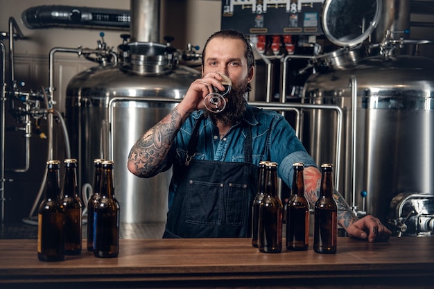 Retrato de fabricante masculino hipster tatuado e barbudo, degustação de cerveja na microcervejaria.