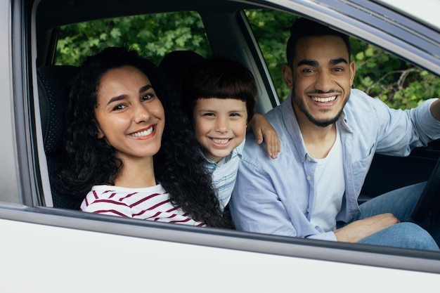 Retrato de família árabe feliz andando de carro juntos, pais alegres do Oriente Médio com filho pequeno sentado no veículo moderno e sorrindo para a câmera pela janela, aproveitando a viagem de verão, closeup