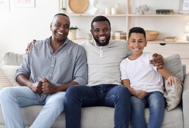 Retrato de família de homens multigeracionais negros felizes posando no sofá em casa, abraçando e sorrindo para a câmera, espaço livre