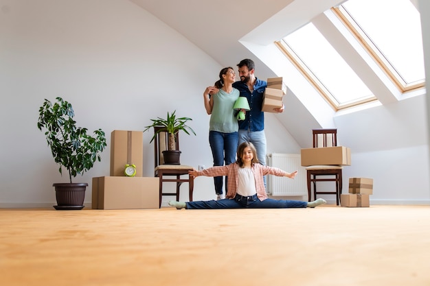 Retrato de família feliz caucasiana, mudando-se para o novo apartamento com uma menina flexível, fazendo exercícios de ginástica e pais carregando pertences.