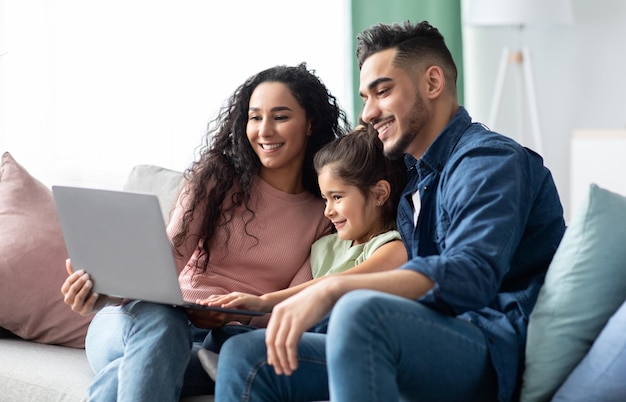 Retrato de família feliz do Oriente Médio usando laptop juntos em casa, pais árabes e sua filhinha bonita relaxando com o computador no sofá na sala de estar, assistindo filmes, curtindo o fim de semana