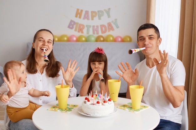 Retrato de família feliz e encantada sentada à mesa e comemorando aniversário olhando para a câmera com expressões positivas acenando com as mãos e soprando o cone de festa