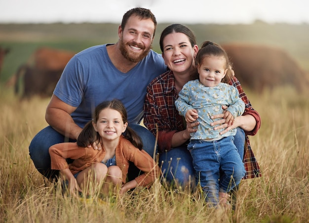 Retrato de família feliz em um campo de fazenda rural com vacas ao fundo Pais de agricultores se relacionando com crianças em um negócio de gado agrícola sustentável com um sorriso e felicidade juntos