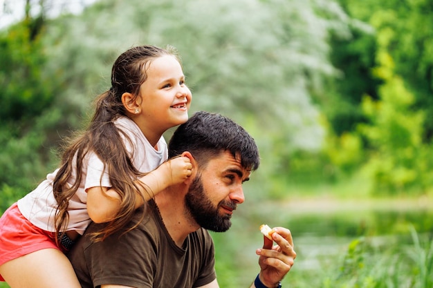 Retrato de família feliz sentado no piquenique na floresta do parque ao redor de arbustos de árvores Filha sentada nas costas dos pais