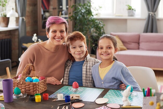 Retrato de família feliz sorrindo para a câmera enquanto está sentado à mesa e pintando ovos para as férias juntos em casa