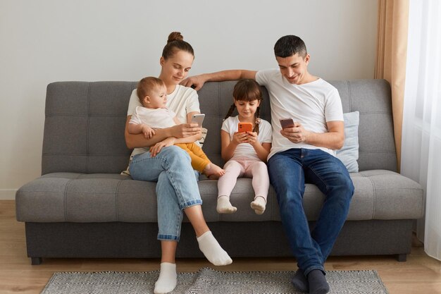 Retrato de família sentada no sofá na sala de estar usando telefones celulares verificando redes sociais parece calma vestindo roupas casuais mãe segurando bebê infantil de joelhos