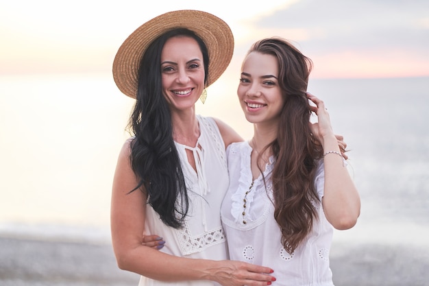 Retrato de feliz atraente sorridente abraçando a filha e a mãe em vestidos brancos no verão ao pôr do sol à beira-mar