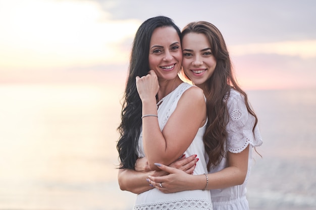 Retrato de feliz atraente sorrindo alegre abraçando filha com mãe em vestidos brancos no verão ao pôr do sol à beira-mar