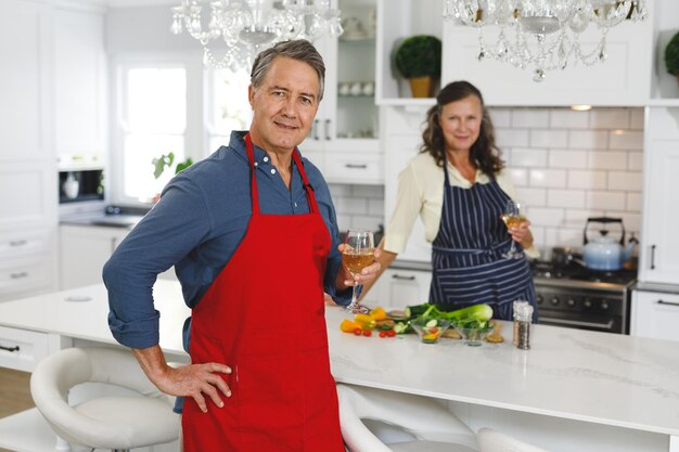 Retrato de feliz casal caucasiano sênior na cozinha cozinhando juntos, bebendo vinho