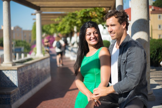 Retrato de feliz casal multiétnico curtindo férias na cidade. Homem sorridente e boa mulher descansando no beco de verão com colunas de mãos dadas e conversando. Felicidade, relações, conceito de viagem