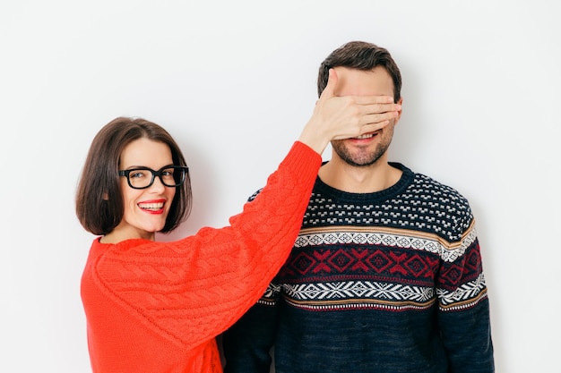 Retrato de feliz cobre os olhos para o marido prepara surpresa para ele usa suéter vermelho casual de grandes dimensões tem sorriso positivo isolado sobre fundo branco vou mostrar algo para você