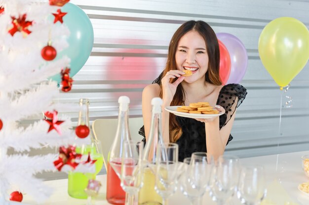 Retrato de feliz e sorridente mulher jovem e bonita asiática segurando e comendo biscoito na festa de natal com comida e bebida