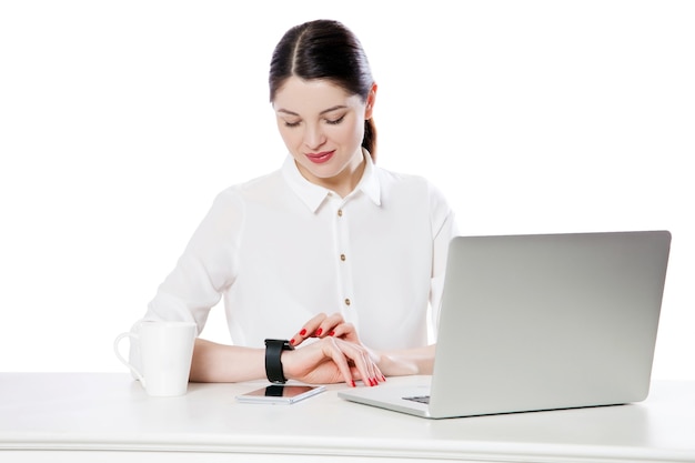 Retrato de feliz empresária morena atraente em camisa branca sentado olhando e tocando seu display smartwatch, lendo algo e sorrindo. tiro do estúdio interno, isolado no fundo branco.